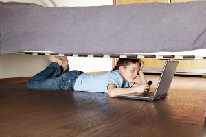 Child with laptop and phone under the bed. Boy playing computer